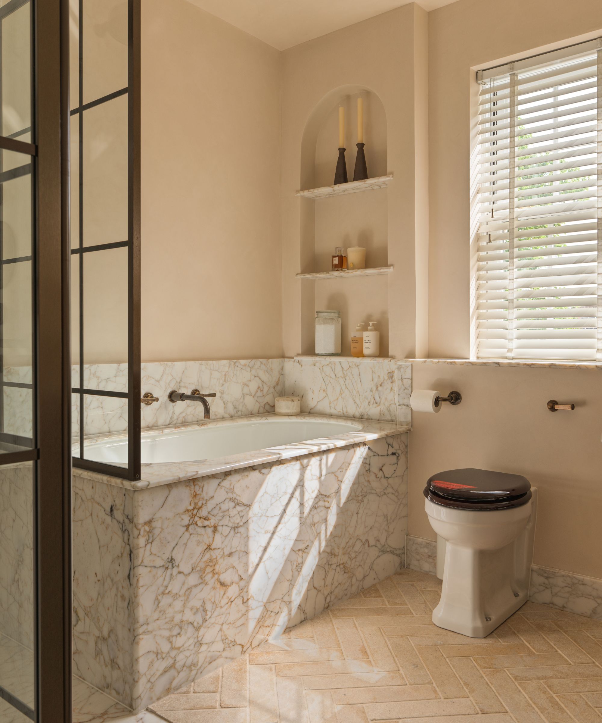 A warm neutral bathroom with terracotta floor tiles and a marble framed bathtub