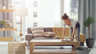 woman tidying up items on coffee table in modern living room, with large gray sofa, big window behind, and a lamp