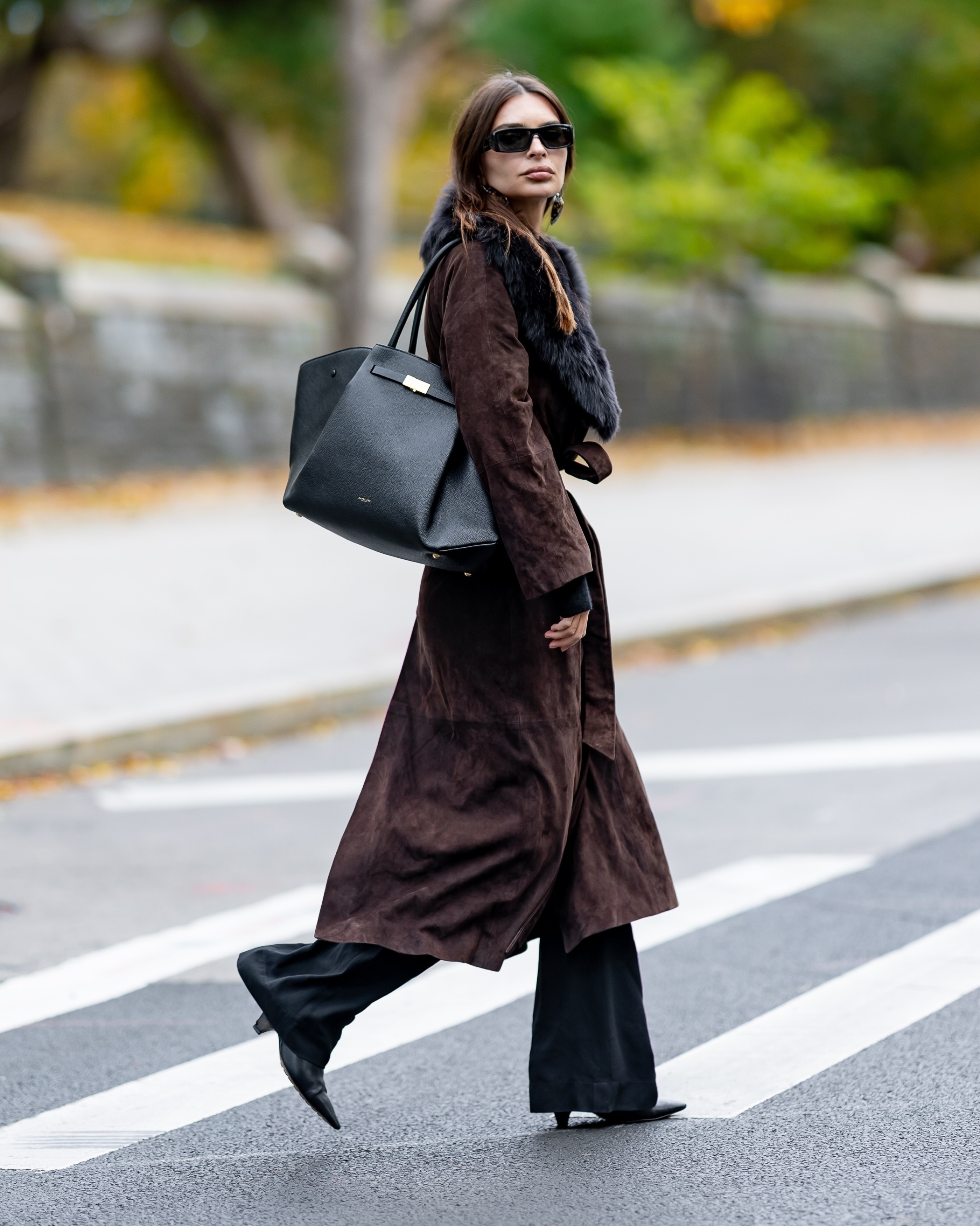 Emily Ratajkowski wears a brown suede trench coat with a fur collar. She styles her coat with black trousers and black kitten heel boots.