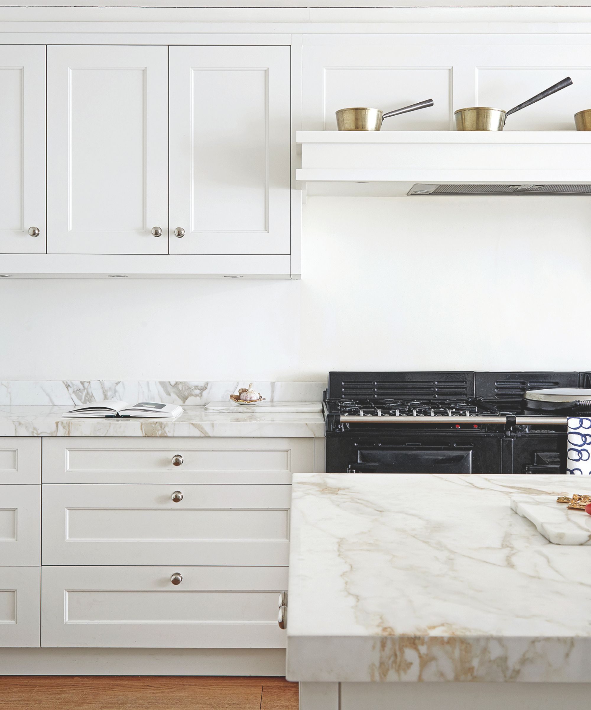 White kitchen with marble countertops and white cabinets, and a large black oven. Above the oven is a white shelf with matching metal pans on top, and to the left of the oven is an open recipe book. The floor is wood.
