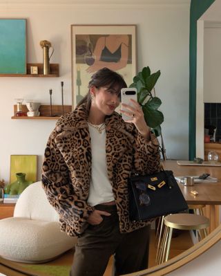 Woman taking mirror selfie while wearing a white shirt, black jeans, a black handbag, and a cheetah-print coat.