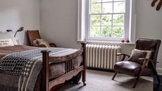 A white bedroom with a metal radiator beneath the window. A rustic wooden bed frame with brown bedding, and a brown leather arm chair opposite. 