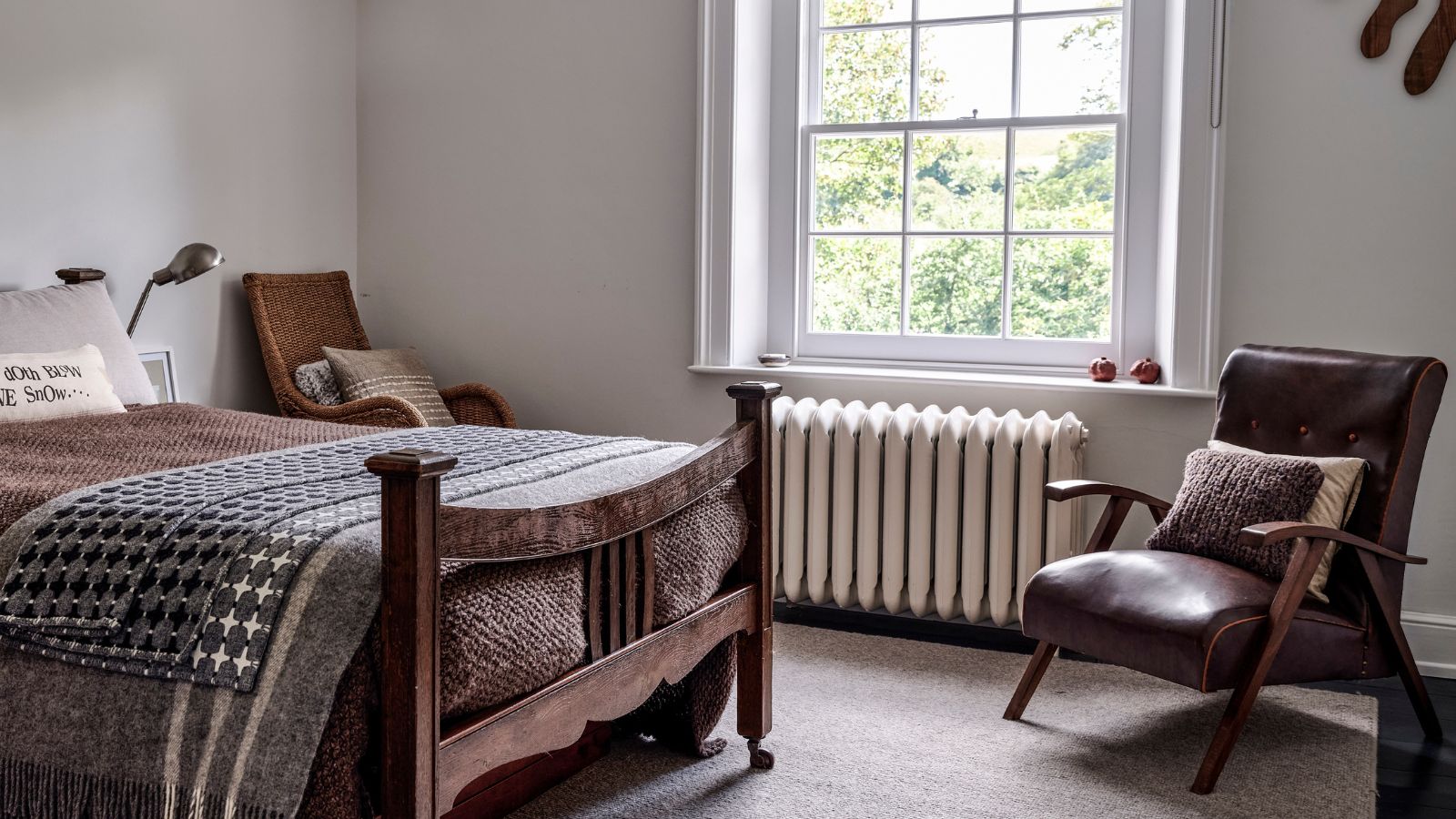 A white bedroom with a metal radiator beneath the window. A rustic wooden bed frame with brown bedding, and a brown leather arm chair opposite. 