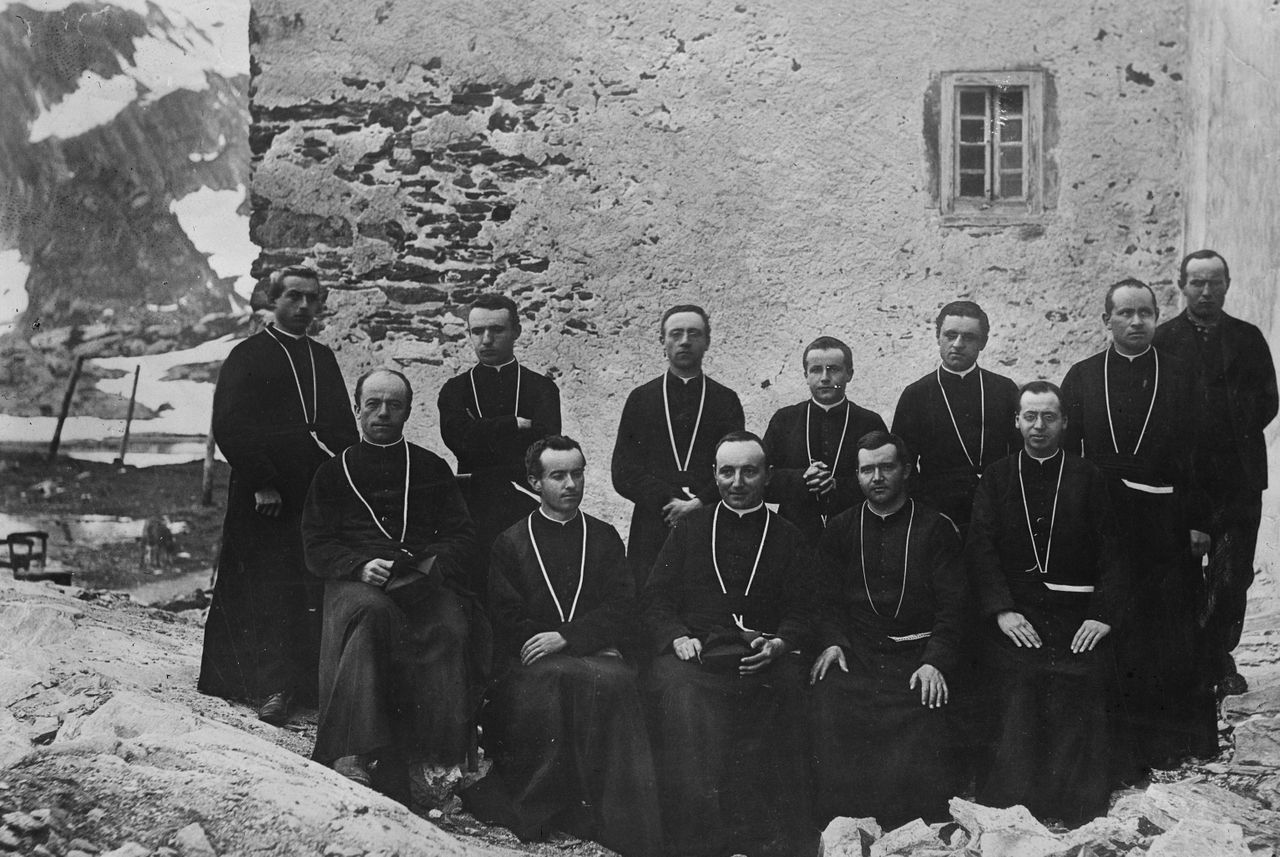 A group of monks wearing long dark robes pose for a formal photograph against an exterior stone wall in a snowy mountain setting.