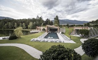 Alternative view of the Seehof Nature Retreat featuring a swimming pool, sun loungers, pathway, lake and surrounding greenery under a cloudy sky