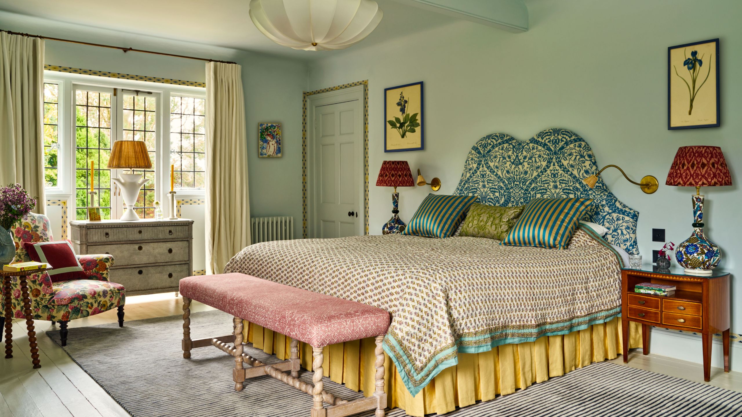 Bedroom painted in Farrow &amp; Ball's Borrowed Light, a blue upholstered headboard, a yellow skirt, a pink bench, large windows with a dresser and lamp in front, and a floral armchair in the corner.