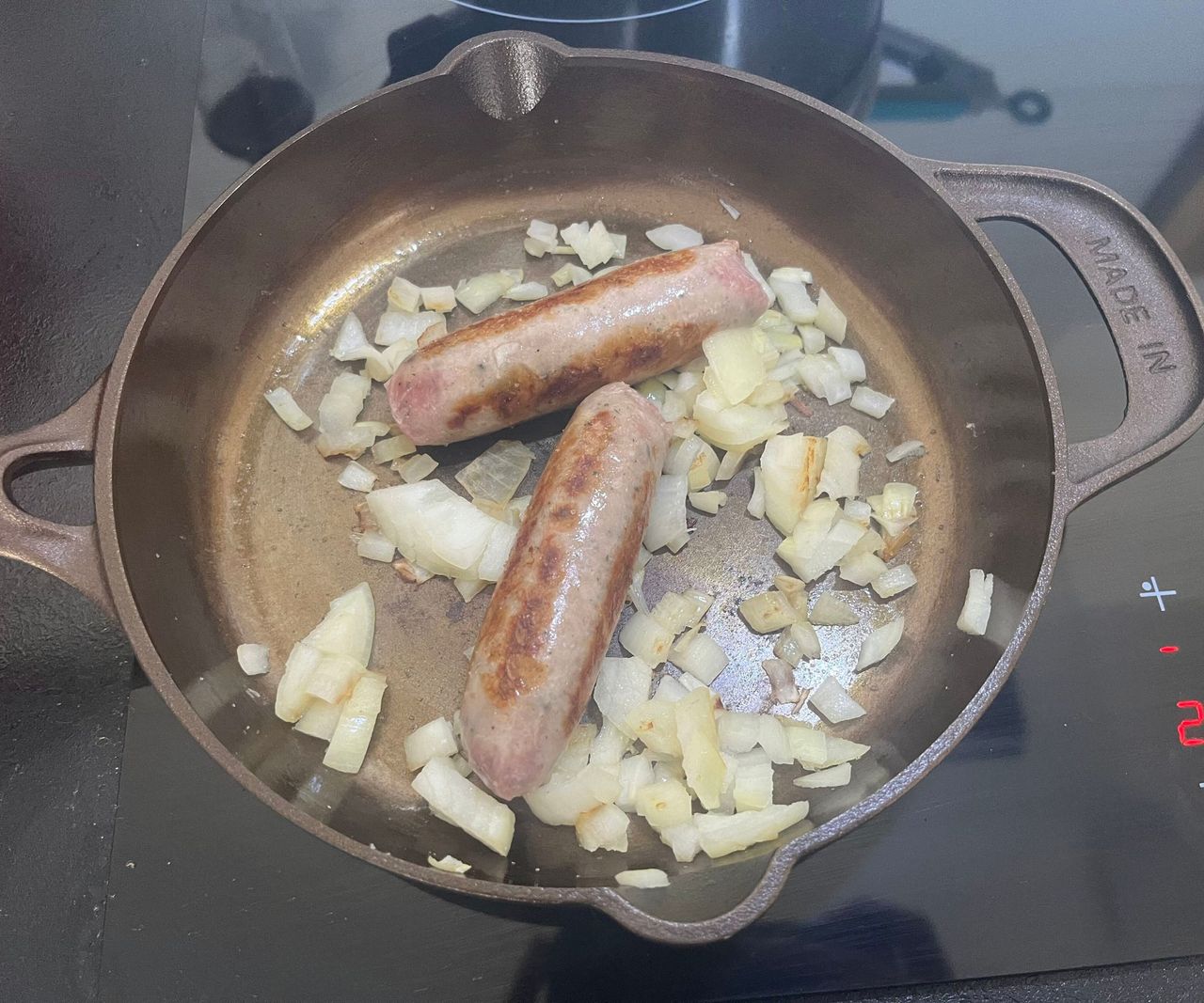 A small bronze colored cast-iron skillet with two sausages and diced onion inside.