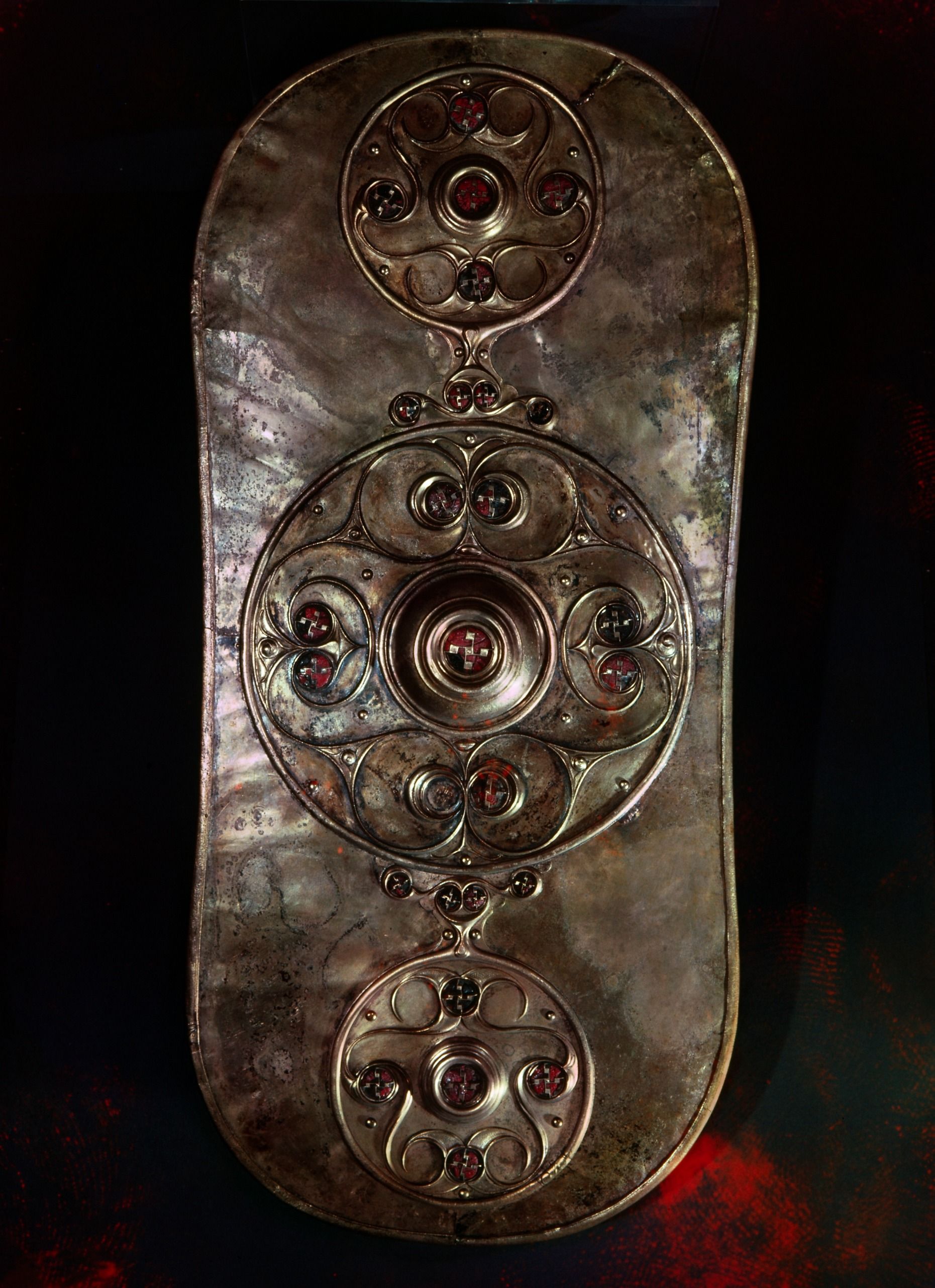 a tall bronze shield with inset red roundel decorations against a black background