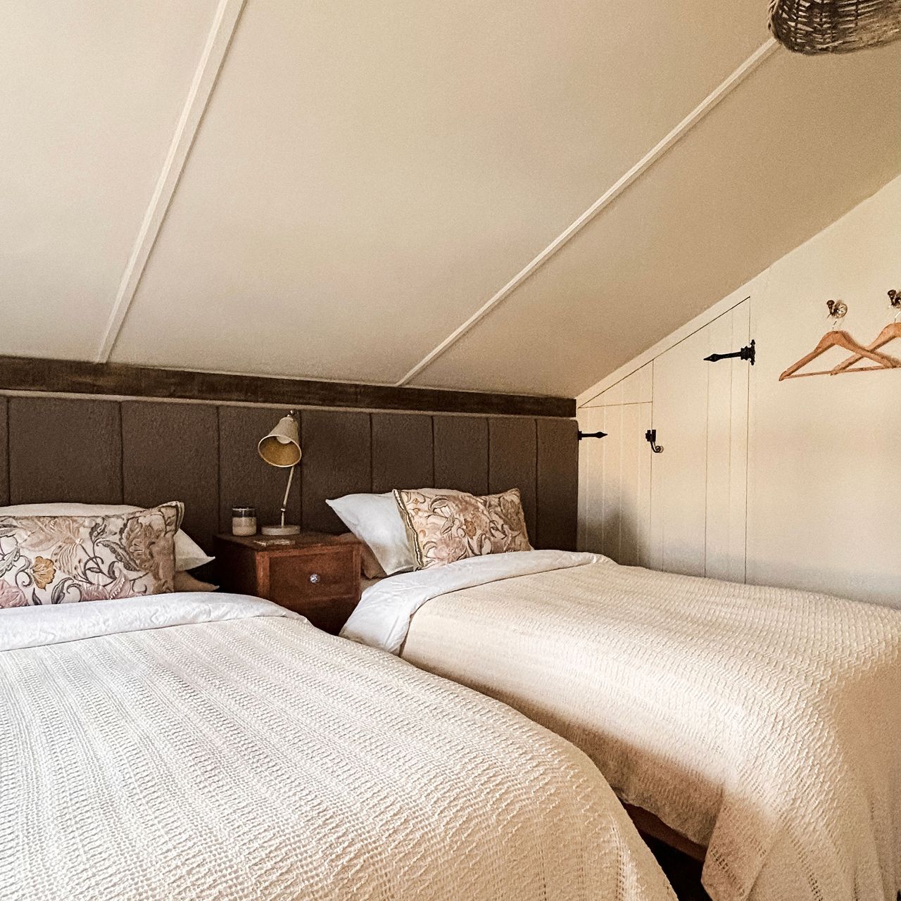 Twin beds in eaves country bedroom with long panelled upholstered headboard behind and wooden bedside table with lamp in between