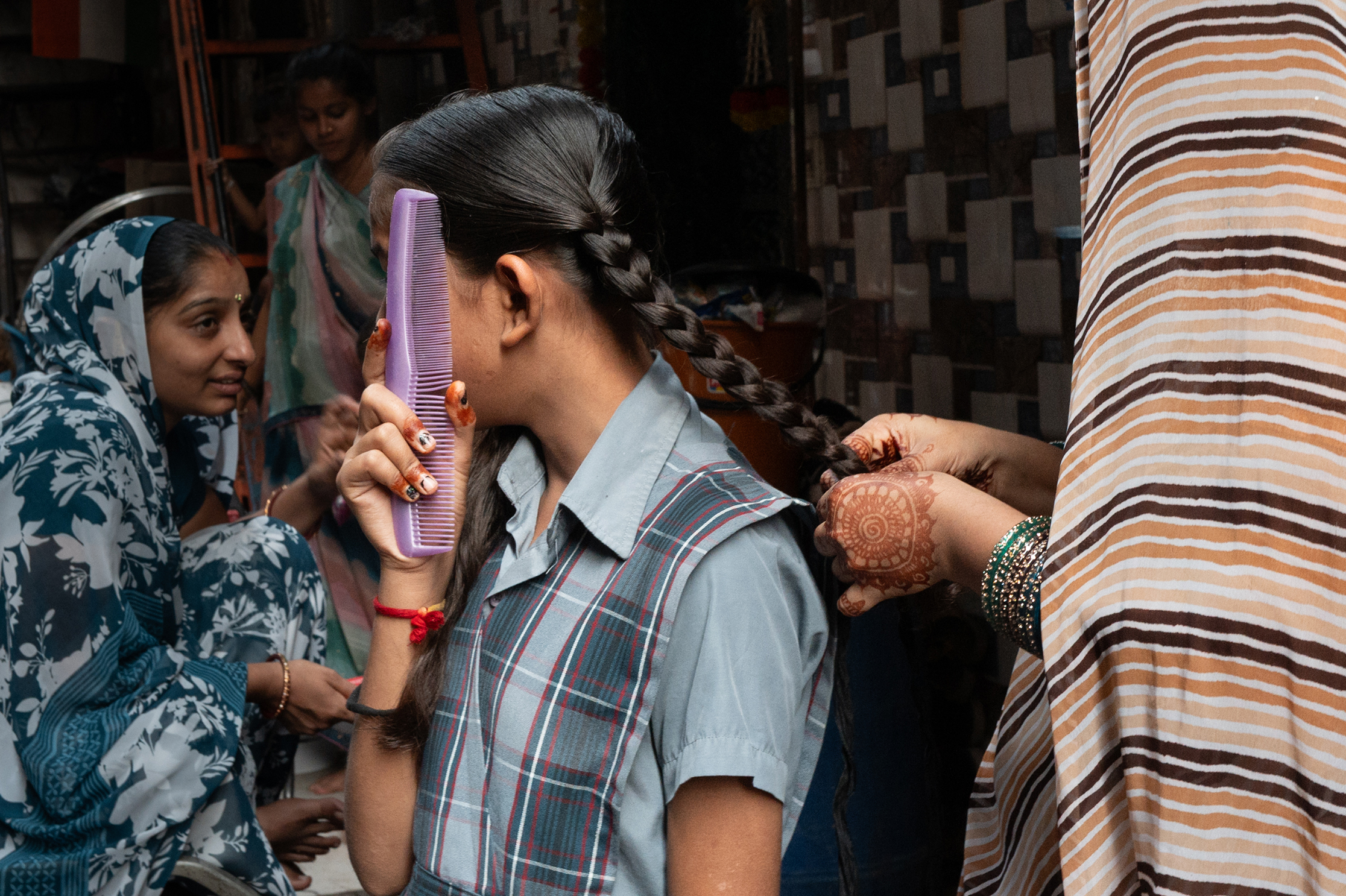 Women gather indoors as one woman combs another&amp;rsquo;s braided hair, surrounded by others in colorful traditional clothing
