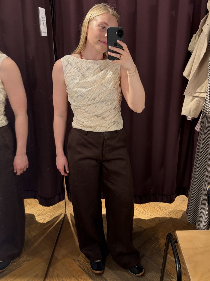 Woman wears draped top, tailored brown linen trousers, trainers