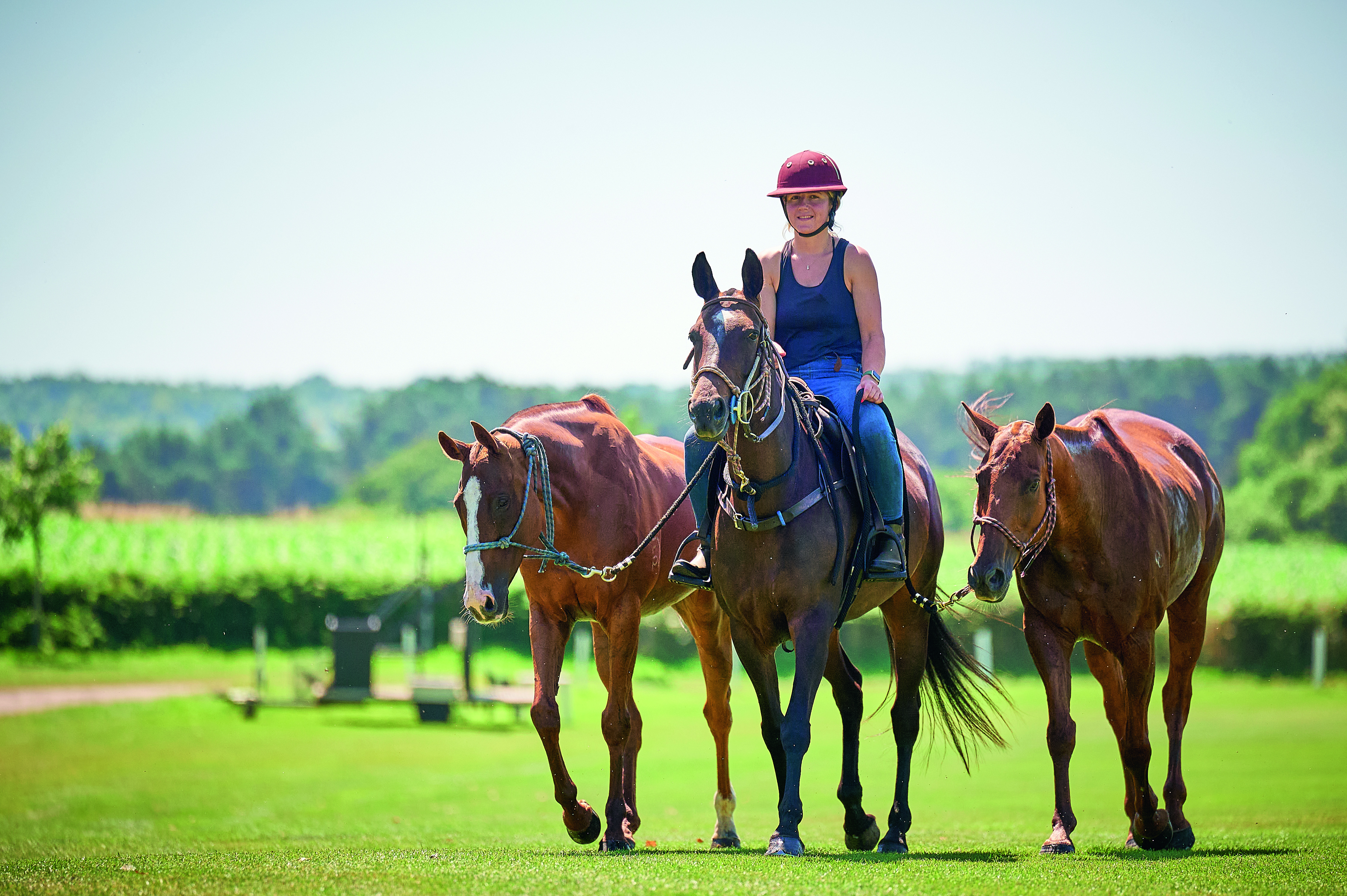 A young woman, Martha Emeney, rides a horse and leads two others across a green field.