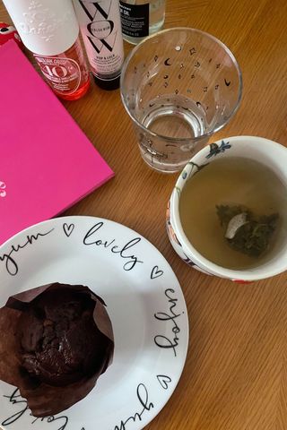 A mug of green tea next to a plate with a chocolate muffin and glass of water