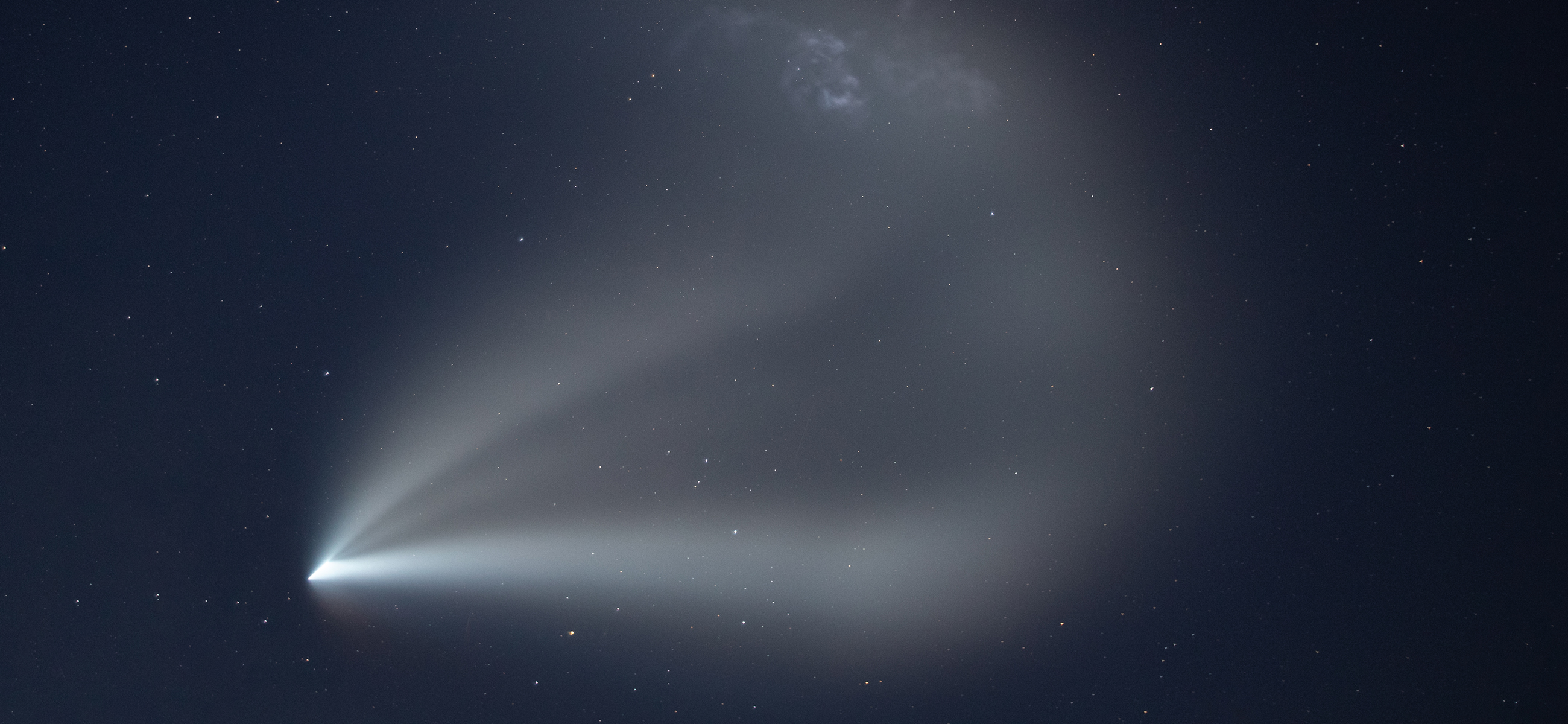 An aurora of white spread out from a pinpoint in the sky like mist from a hose, highlighting the rocket's expanding plume as it climbs the atmosphere against a dark, starry night sky.