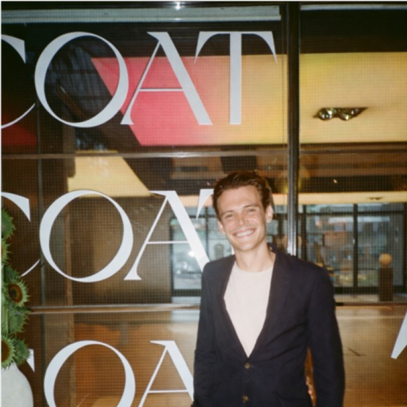 Image of a smiling man in a blazer and white shirt standing in front of a window that features the word Coat in large format lettering