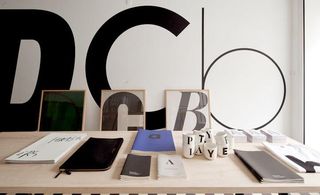 A light wooden table displaying a series of exercise books of differing sizes. In the background there are large scale black letters displayed on a white wall.