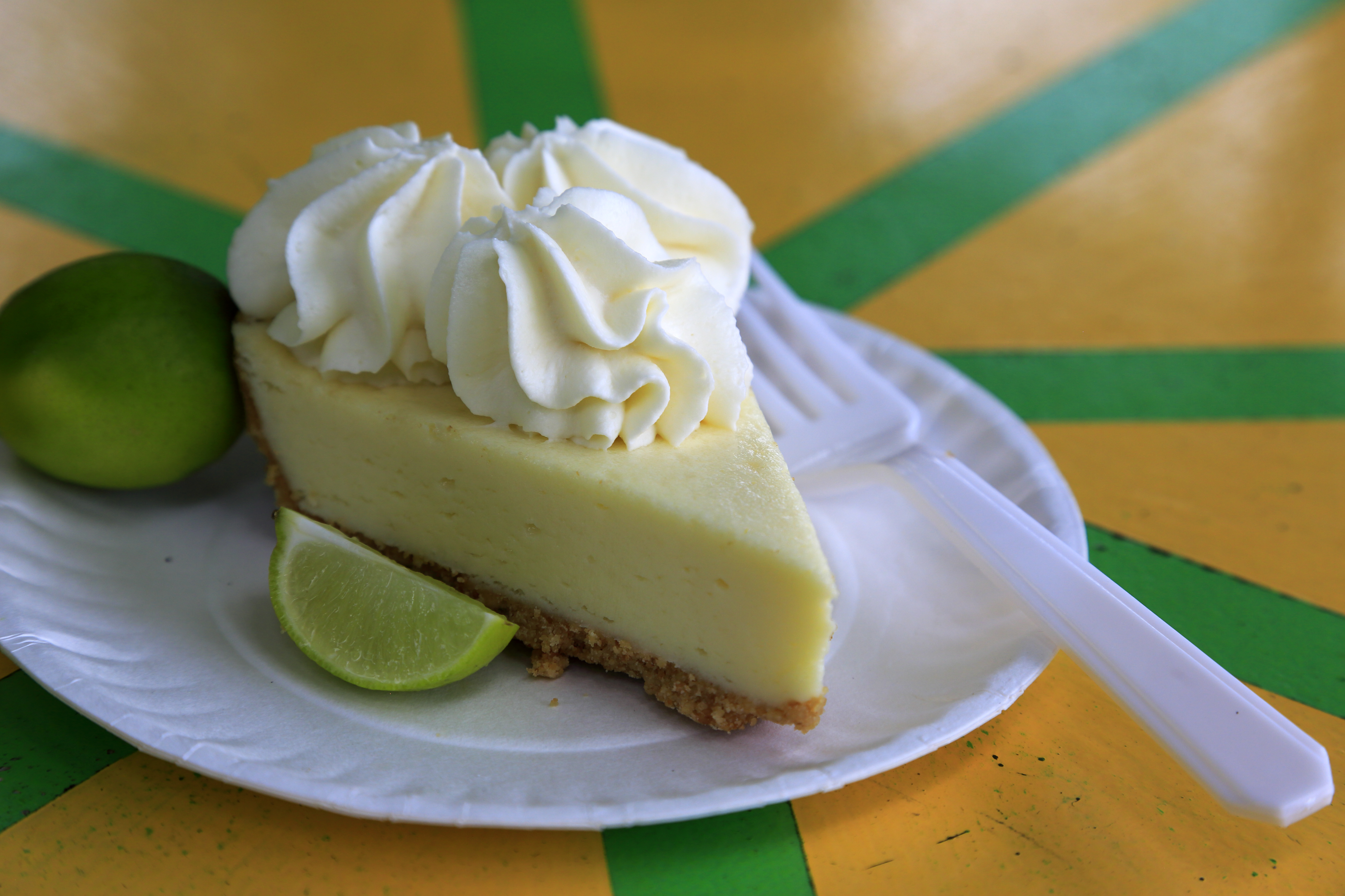 A slice of Key lime pie on a white paper plate with a white fork and lime wedges