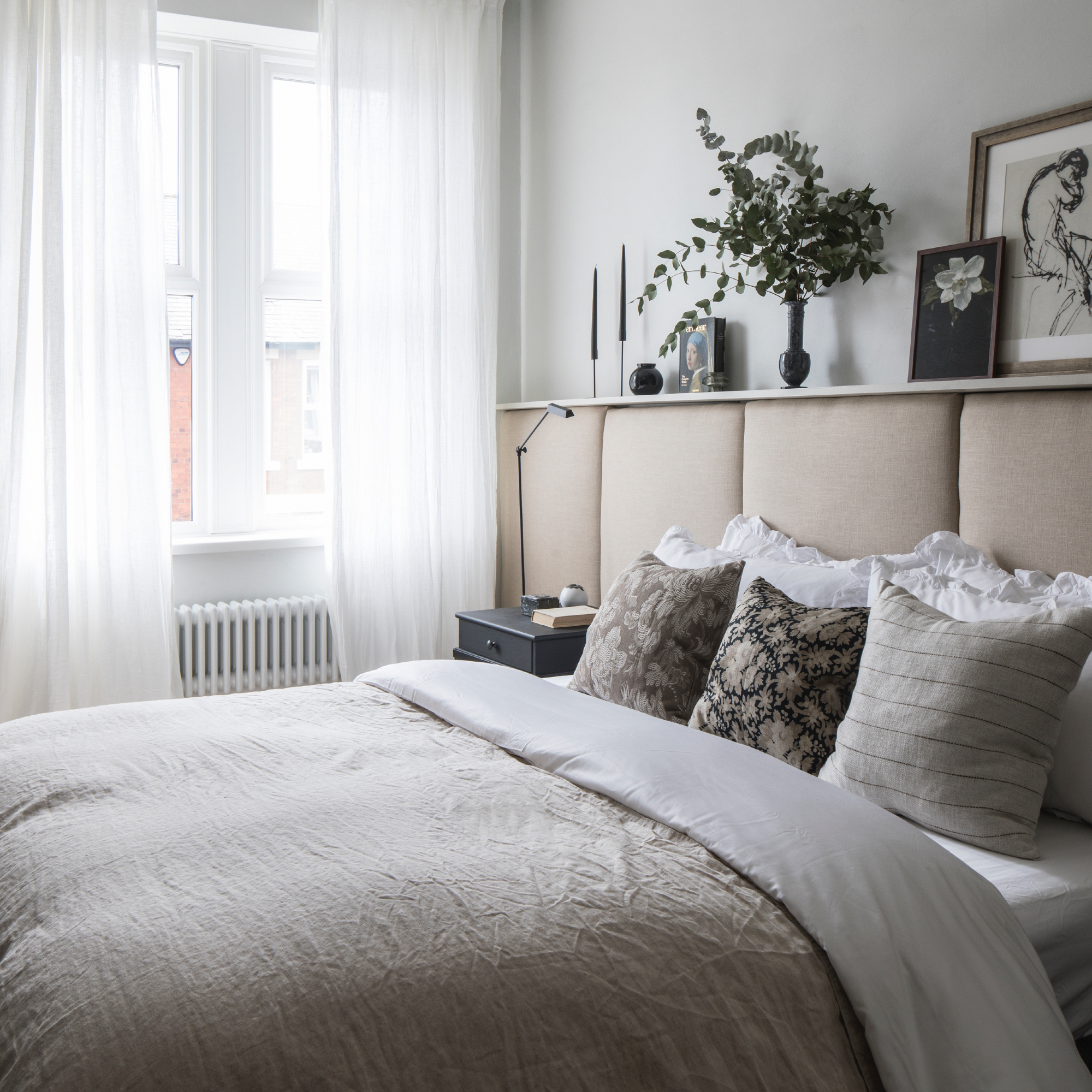 bedroom curtain mistakes, neutral bedroom with padded oatmeal headboard, taupe bedding, patterned cushions, shelf above headboard, vintage chair in corner, white voile curtains, radiator, wooden floor boards, artwork