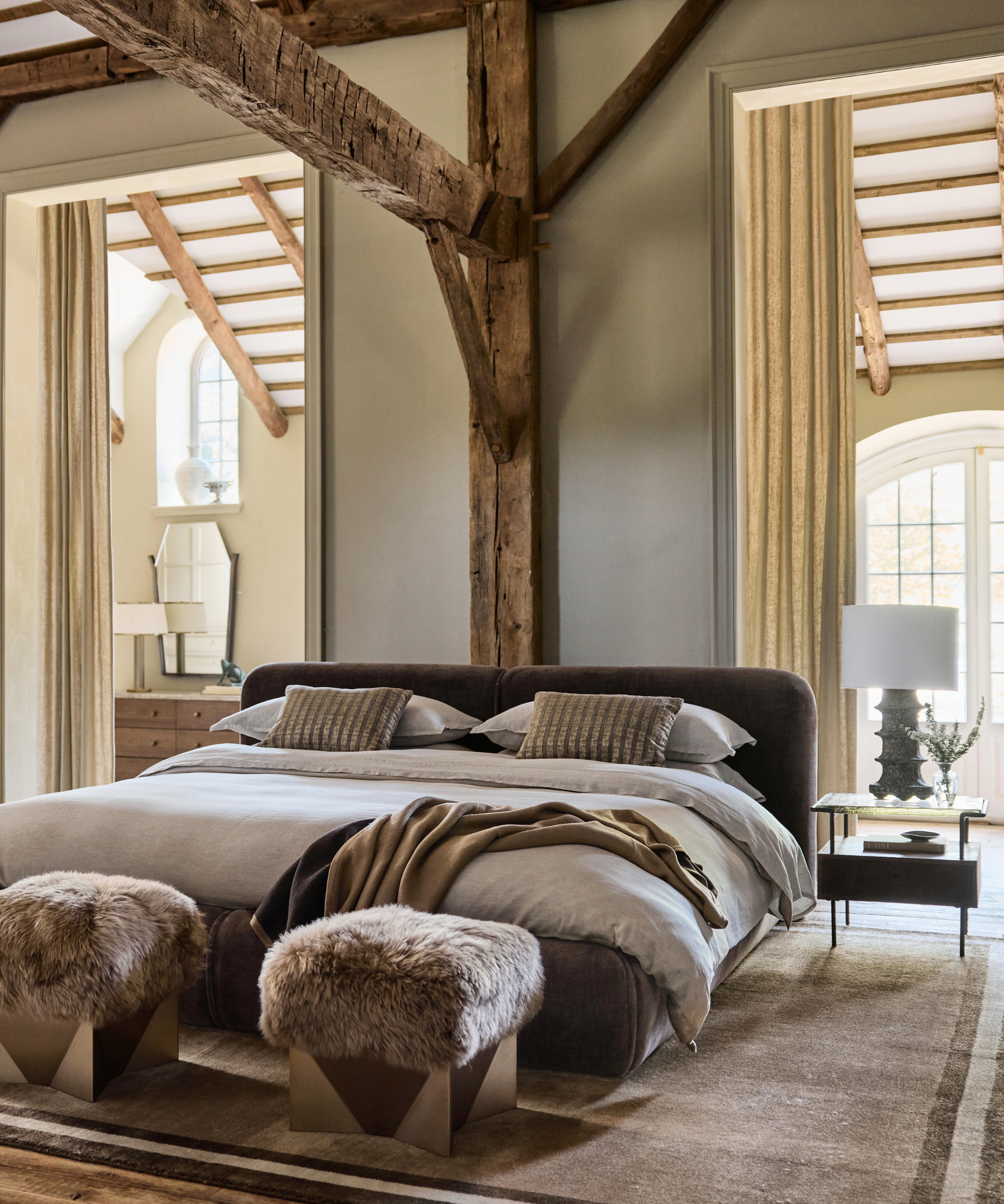 a bedroom with exposed wood beams, a large bed with gray sheets, two unique ottomans, and a bedside table with a lamp