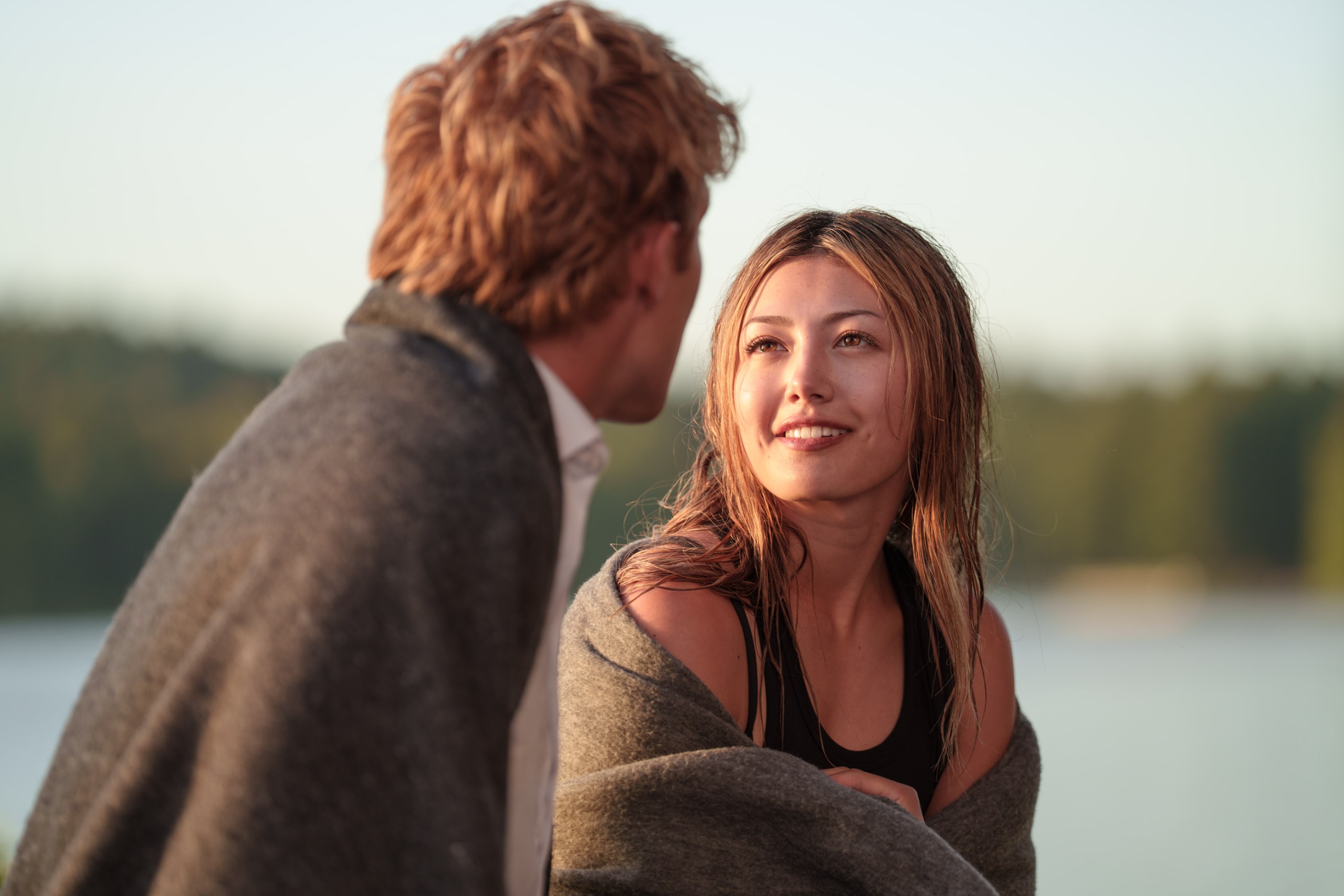 A teenage girl (Miku Martineau as Teagan Tao) with a towel drapped around her shoulders sits with a boy in front of a lake, in Netflix's 'Crew Girl'.