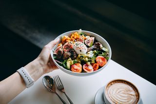 Close up of woman's hand holding a bowl of fresh beef cobb salad, serving on the dining table. Ready to enjoy her healthy and nutritious lunch with coffee. Maintaining a healthy and well-balanced diet. Healthy eating lifestyle