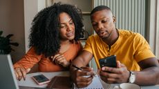 A young couple look at their phones while financial planning at their dining room table.
