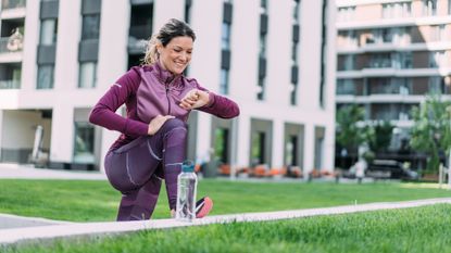 Woman in tracksuit and sports leggings looks at her watch. She is outside, with one foot on a raised ledge, next to a water bottle