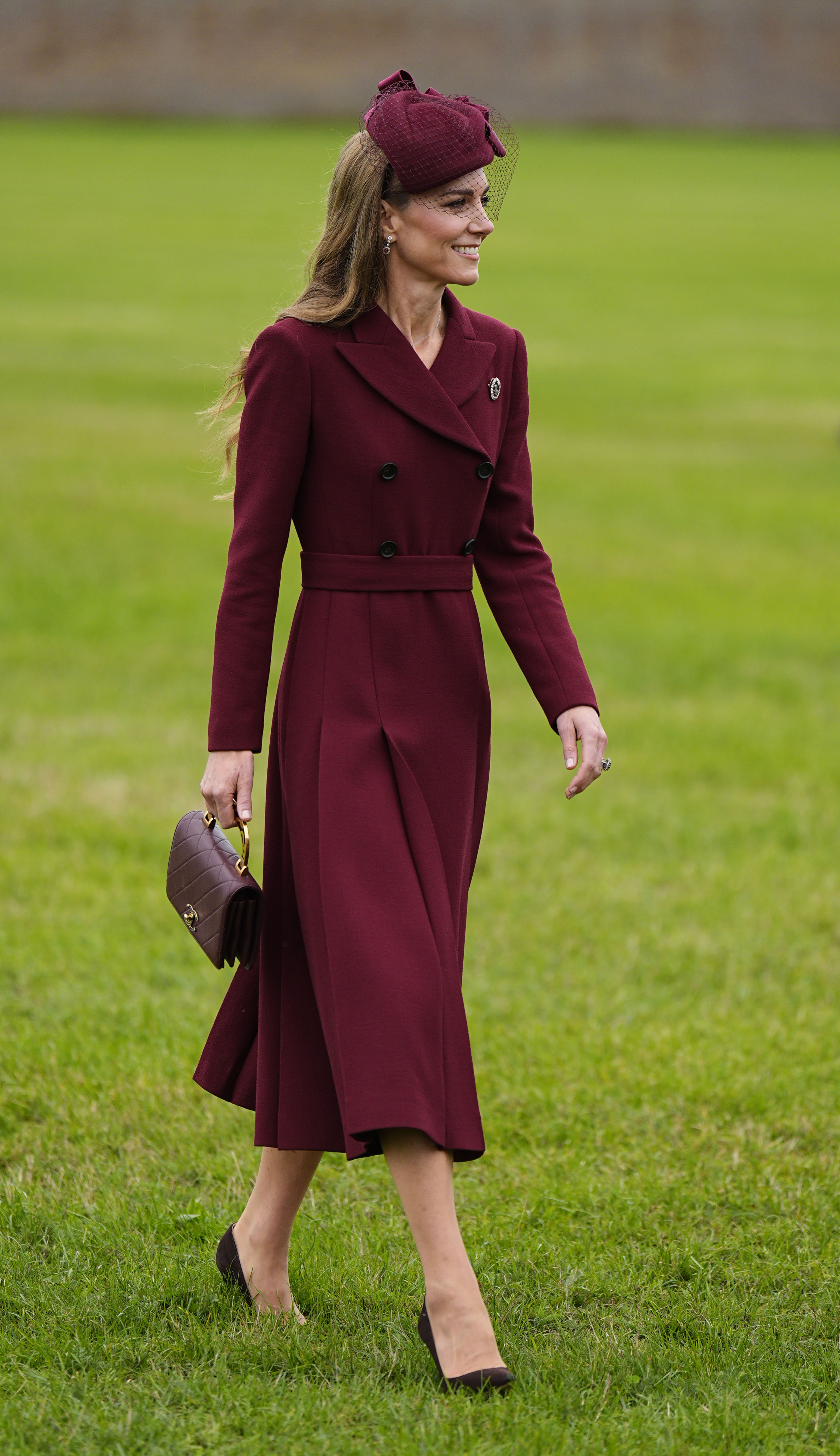 Kate Middleton walks outside on grass wearing a burgundy coat with chocolate brown heels. She carries a burgundy Chanel bag.