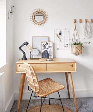A white home office with a light wooden desk with a lamp, mirror and prints on it, a rattan chair in front of it, a circular mirror, white grid organizer and wooden pegs with a plant in a woven basket hung on the wall
