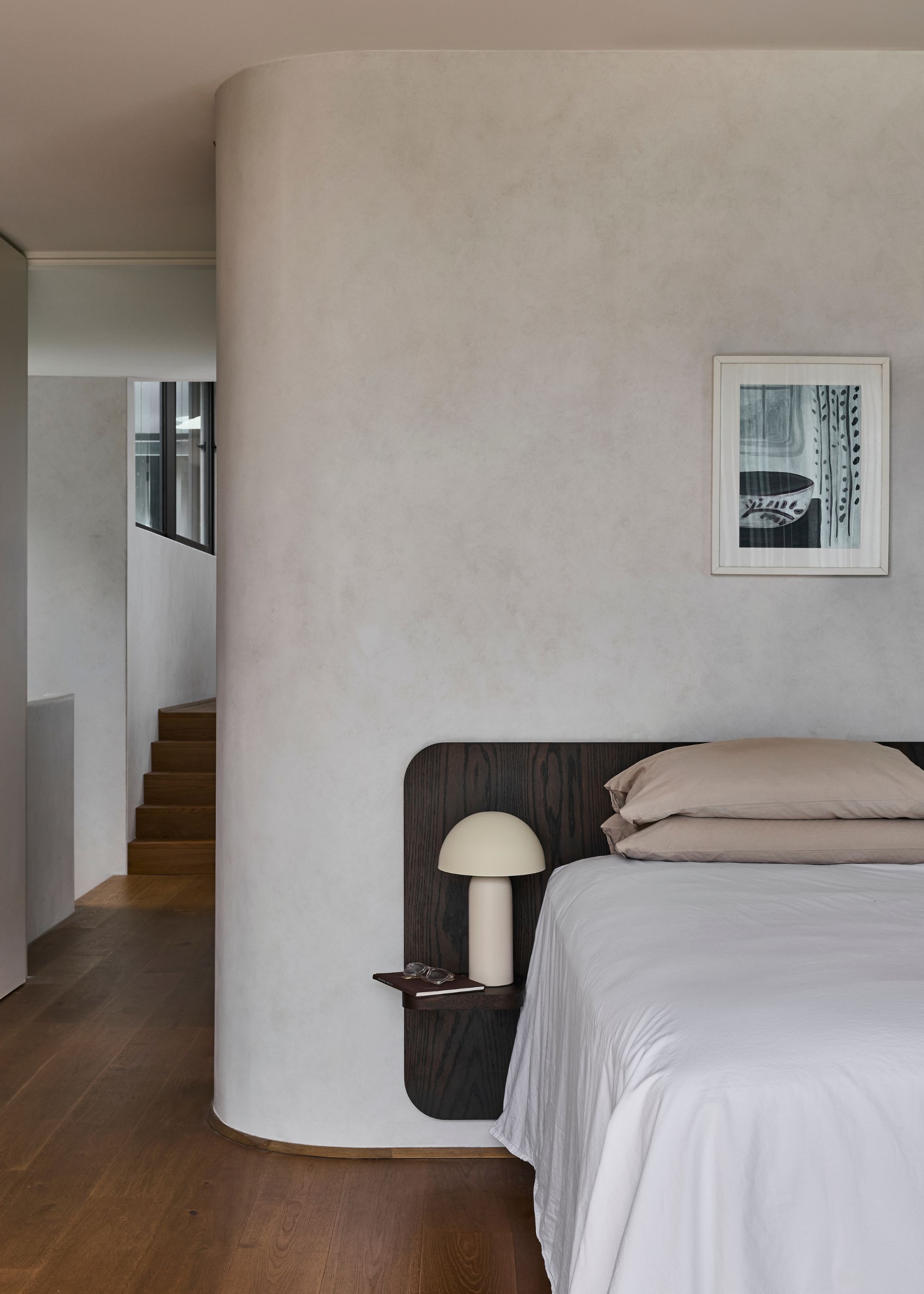 An off-white bedroom with textured curved walls, a wood headboard, white bedding, a mushroom lamp, and a framed print