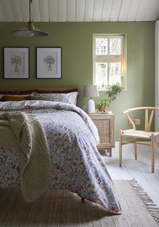 green farmhouse style bedroom with floral bedding, artwork, wooden chair, side table, white floor boards, shiplap ceiling and rise and fall pendant