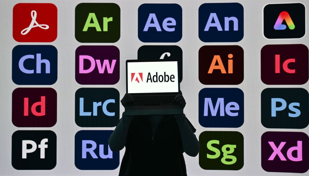 In this photo illustration, logos of Adobe Apps are displayed on screens with a shadowed person holding a laptop that reads Adobe