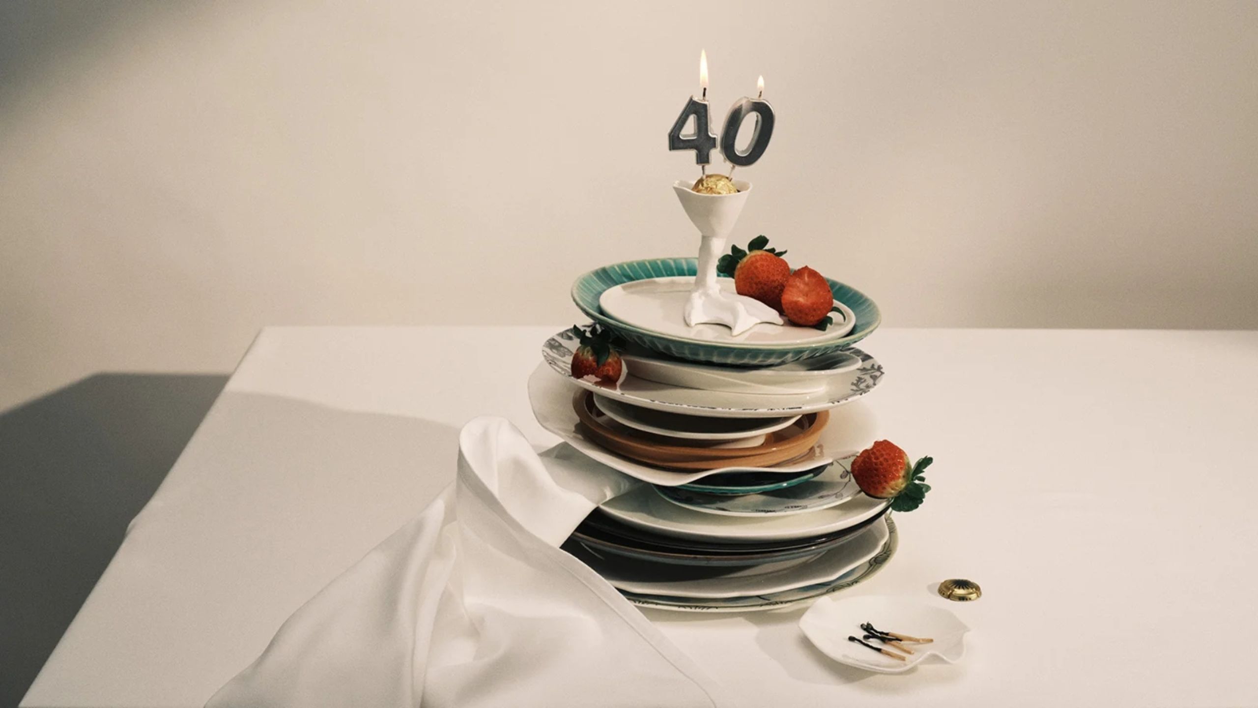 A pile of serax tableware on a white tablecloth with a small egg-size cup with a 40 candle lit on top and a small tray of used match sticks on the side