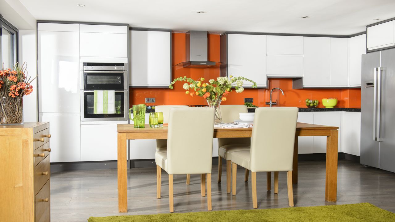 wooden dining table with light coloured chairs with white cabinets and an orange wall