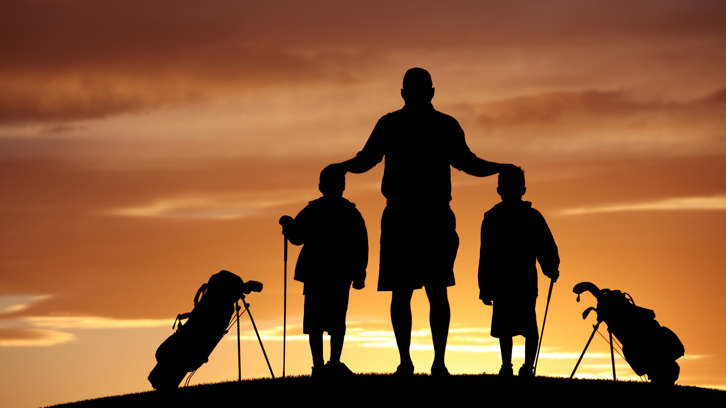 A silhouette of a father with his two sons standing next to their golf bags on the golf course at sunset, with the father placing his hands on the two boys heads in a supportive and encouraging manner.