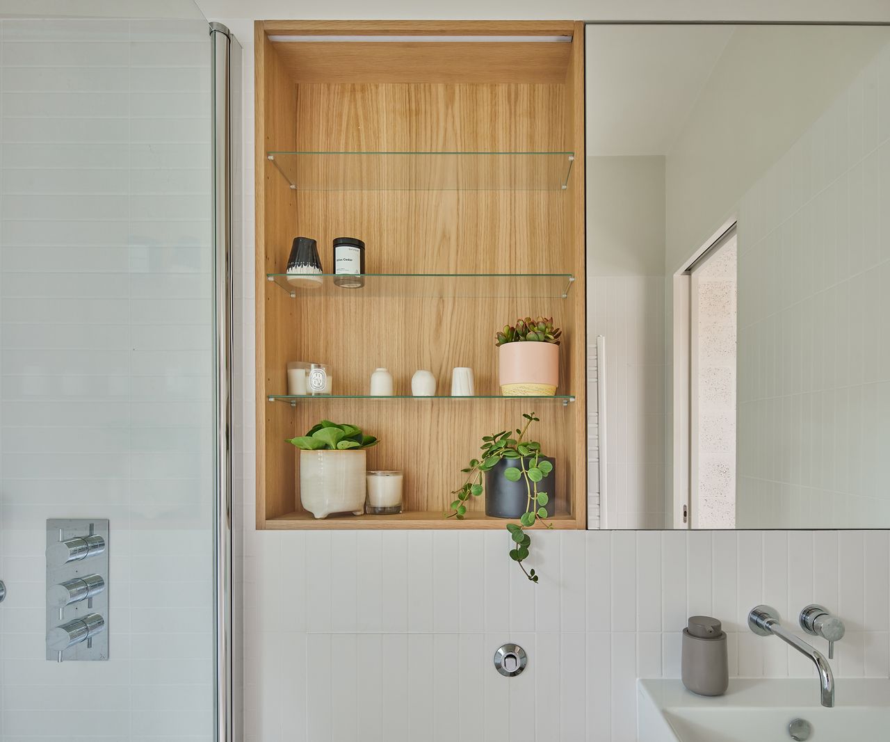 A wood and glass cabinet above the sink in a white tiled contemporary bathroom. There are small potted plants and luxury scented candles on the shelves with a mirror to the right hand side