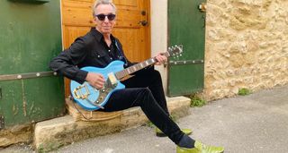 Keith Streng sits on a doorstep wearing all black, dark shades and slime green leather shoes. He blues a blue Supro.
