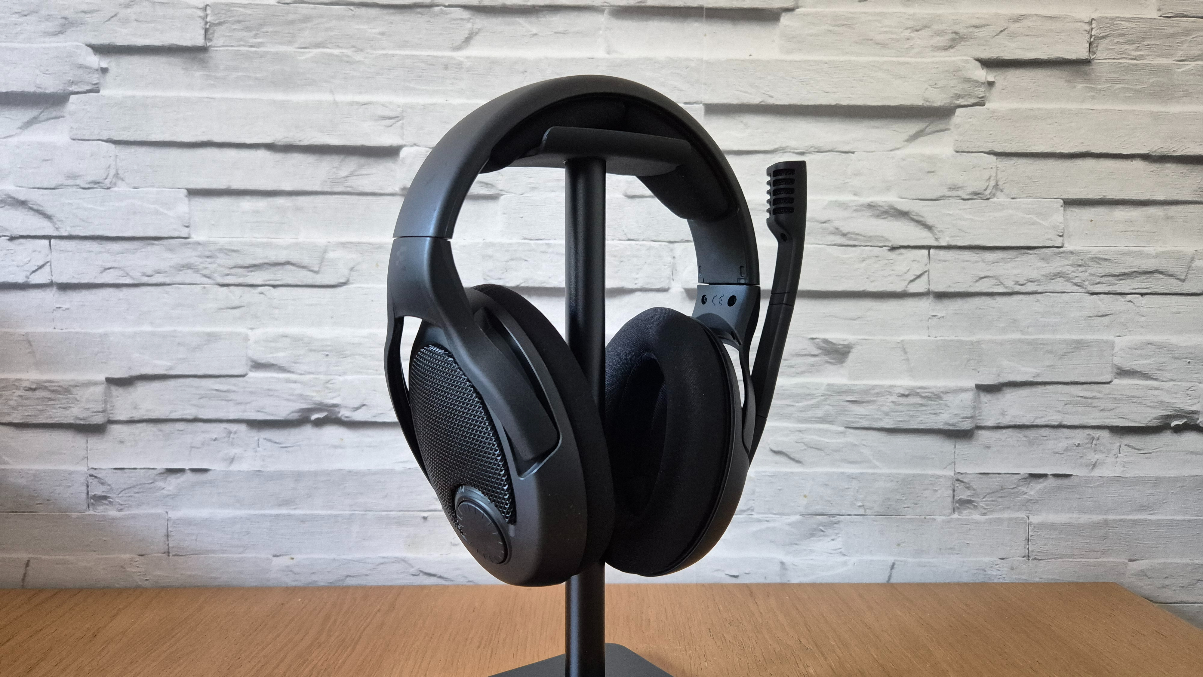 The Drop + Epos PC38X headset on a stand sitting on a wooden surface infront of a white brick background