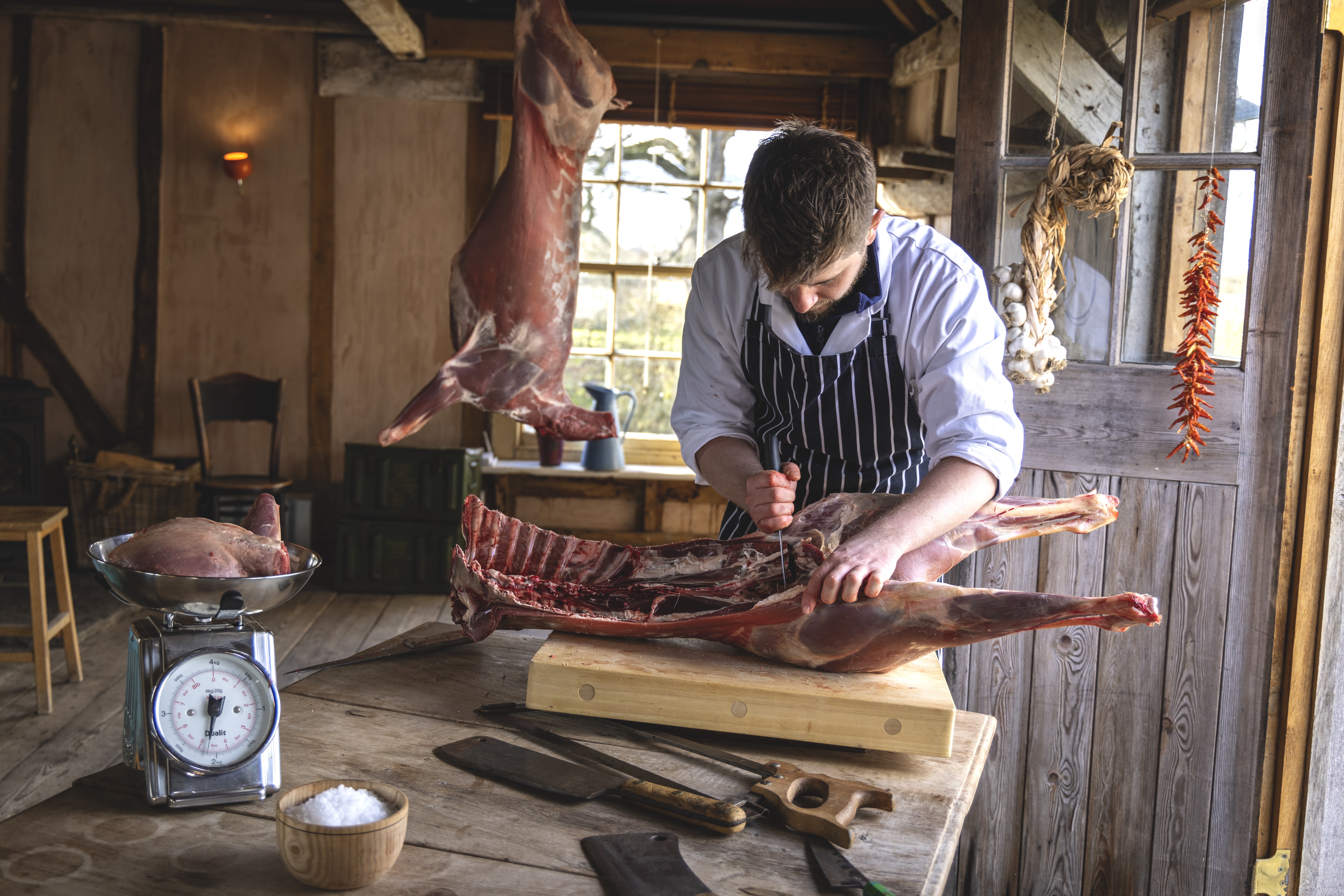 A man in a butcher's uniform and blue and white stripe apron carves up an animal carcass on a wooding chopping block