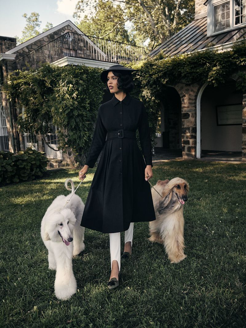 Nara Smith wears volumious black coat with a hat and white leggings while walking two large dogs on grass
