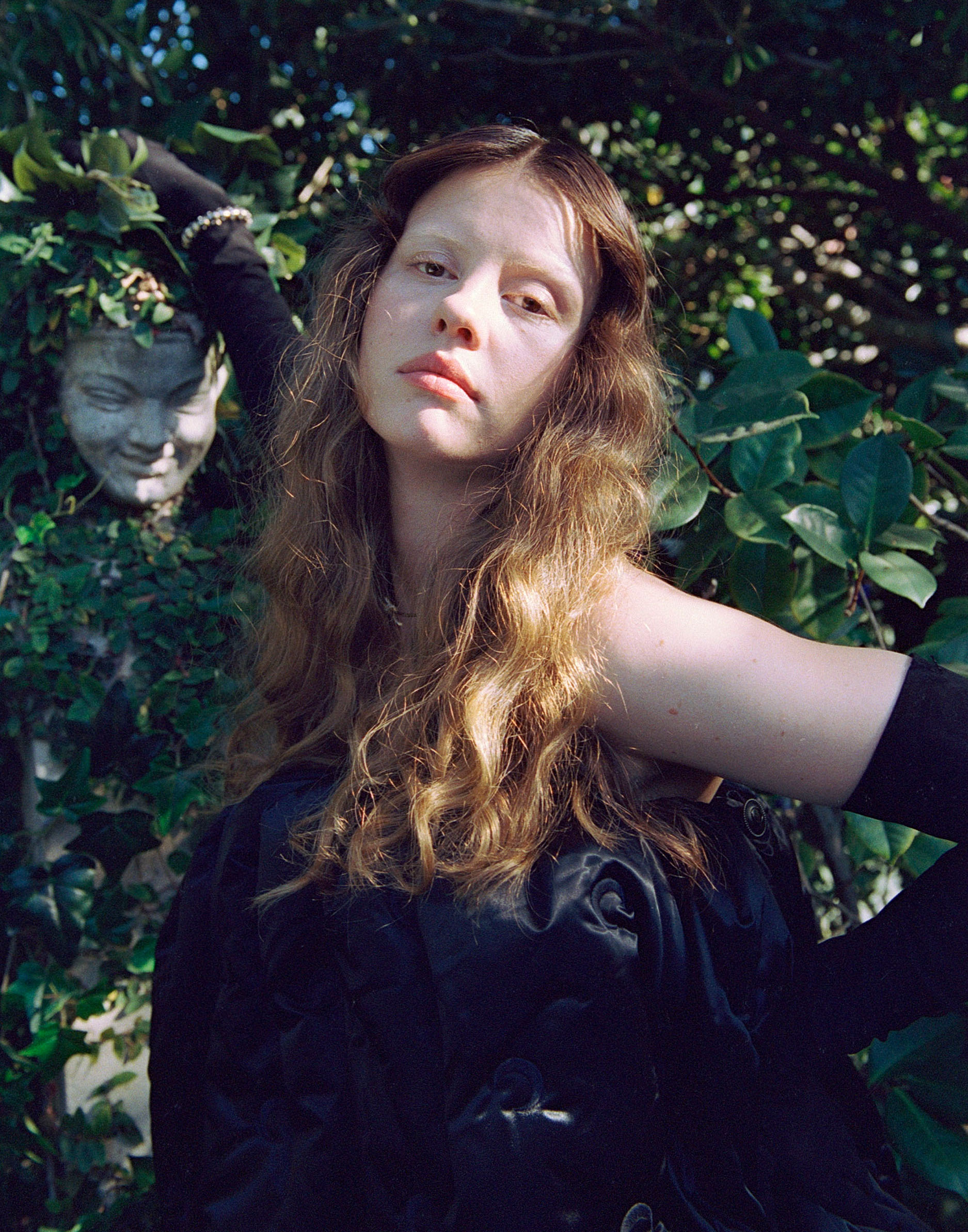 Mia Goth photo shoot for Who What Wear&#039;s October Cover Story. In this image, Mia is standing in front of greenery covered wall. She is wearing a voluminous black dress with black gloves.