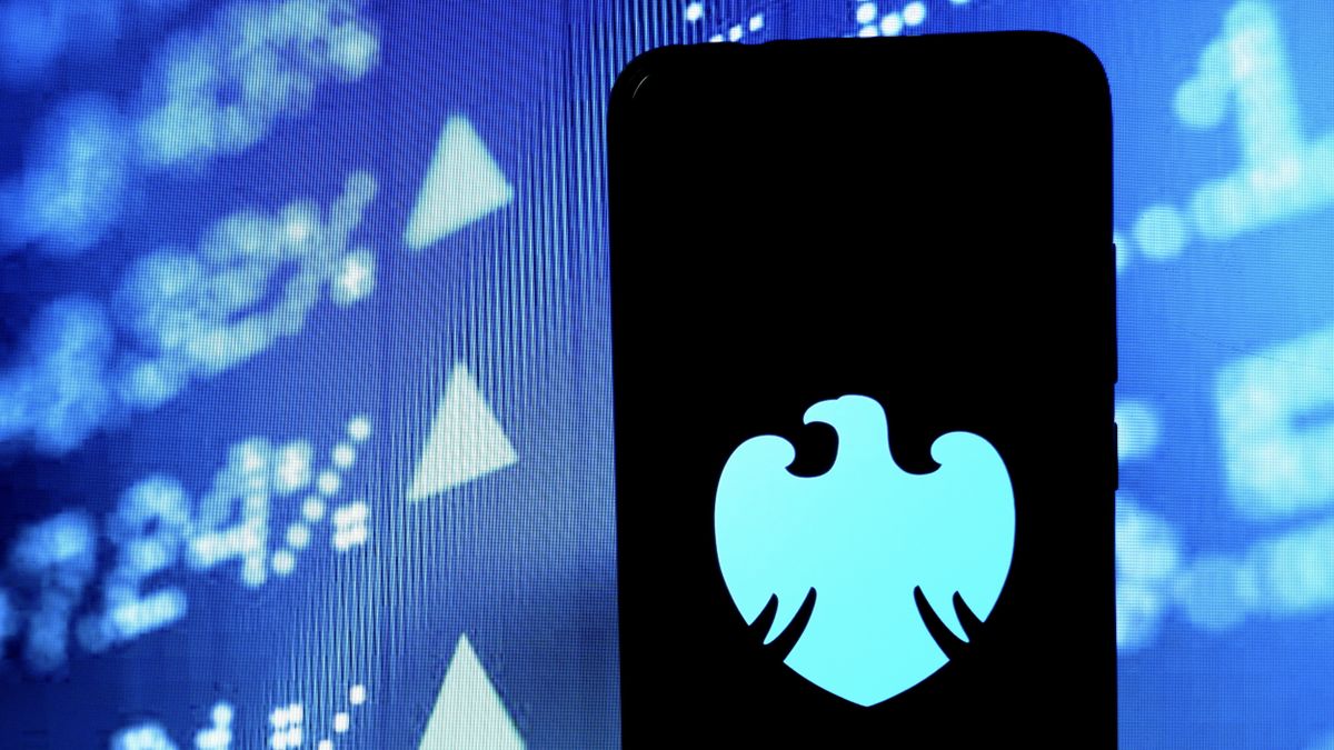 The Barclays logo on a smartphone in front of a digital board with company share prices