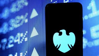 The Barclays logo on a smartphone in front of a digital board with company share prices