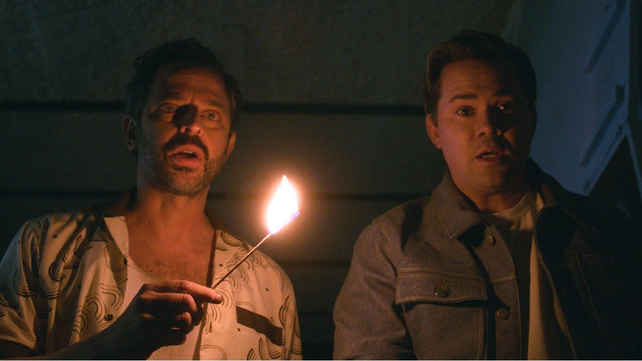 From left to right: Nick Kroll looking shocked and holding a match that's on fire with his left hand and Andrew Rannells looking shocked next to him.