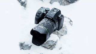 Et sort Pentax K-70 står på en sten i sne med lidt sne på.