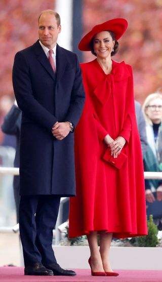 Kate Middleton in a red bow cape coat with Prince William in a blue suit.