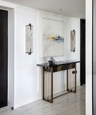 White painted hallway with large rectangular panels, marble flooring, metallic console table with table lamp. marble effect wall lamps, large square artwork. neutral scheme