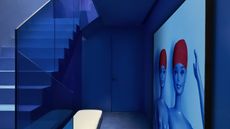 Image of a stairwell and hallway drenched in electric, cobalt blue. The stairwell has blue glass sides and there is a giant all-blue photo of Barbie dolls in red swim caps on the wall.