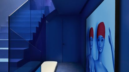 Image of a stairwell and hallway drenched in electric, cobalt blue. The stairwell has blue glass sides and there is a giant all-blue photo of Barbie dolls in red swim caps on the wall.