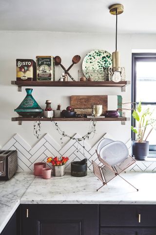 Wooden shelves holding vintage ornaments, house plants and chopping boards with herringbone tiling, in a dark navy kitchen with gold accents and and marble surfaces
